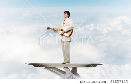 Businessman on metal tray playing acoustic guitar against blue sky background Businessman on metal tray playing acoustic guitar against blue sky background 48857125