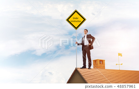 Young businessman on house brick roof holding yellow signboard. Mixed media Young businessman on house brick roof holding yellow signboard. Mixed media 48857961