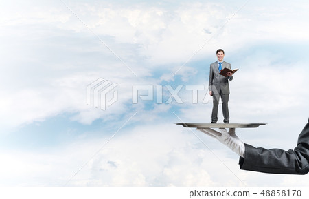Businessman on metal tray with red book in hands against blue sky background Businessman on metal tray with red book in hands against blue sky background 48858170