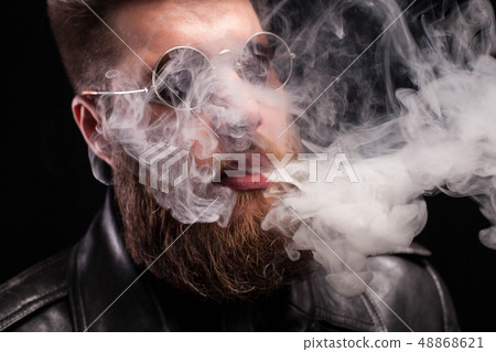 Close up portrait of bearded young man with sunglasses exhaling cigarette smoke over background Close up portrait of bearded young man with sunglasses exhaling cigarette smoke over background 48868621