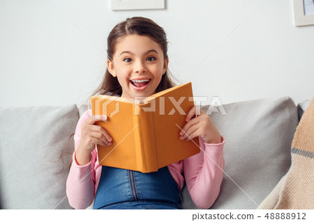 Little girl at home sitting holding book looking camera surprised Little girl at home sitting holding book looking camera surprised 48888912