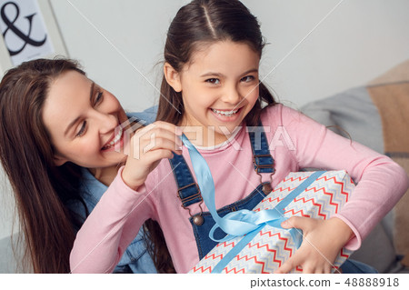 Mother and daughter at home birthday sitting girl opening gift box looking camera cheerful Mother and daughter at home birthday sitting girl opening gift box looking camera cheerful 48888918