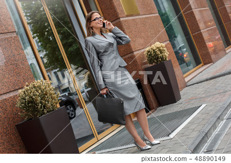 Business woman in eyeglasses standing on the city street with case talking on smartphone joyful 48890149