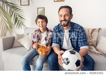 Father and little son at home sitting on sofa boy eating potato chip dad with ball watching football Father and little son at home sitting on sofa boy eating potato chip dad with ball watching football 48891481