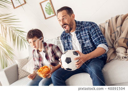 Father and little son at home sitting on sofa boy holding potato chip and dad with ball watching 48891504