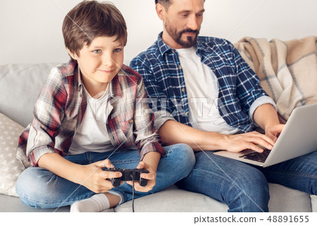 Father and little son at home sitting on sofa dad working on laptop while boy close-up playing game 48891655