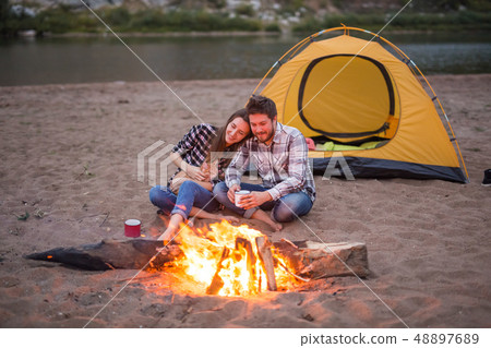 People, tourism and nature concept - Romantic evening, man embrace woman sitting near a campfire People, tourism and nature concept - Romantic evening, man embrace woman sitting near a campfire 48897689