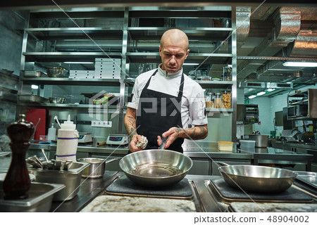 Cooking process. Brutal male chef with several tattoos on his arms holding a frying pan with oil Cooking process. Brutal male chef with several tattoos on his arms holding a frying pan with oil 48904002