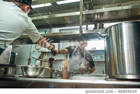 Take it. Young professional chef in apron giving a pepper grinder to his assistant while cooking Take it. Young professional chef in apron giving a pepper grinder to his assistant while cooking 48904039