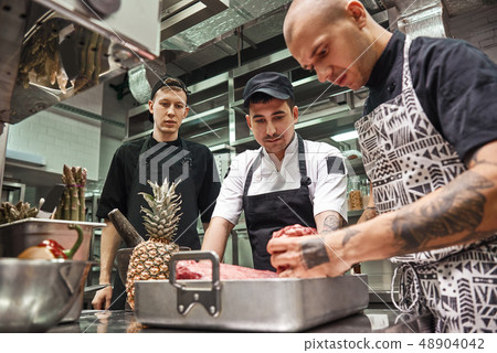 Right choice. Professional chef in apron holding a red meat and teaching his assistants how to Right choice. Professional chef in apron holding a red meat and teaching his assistants how to 48904042