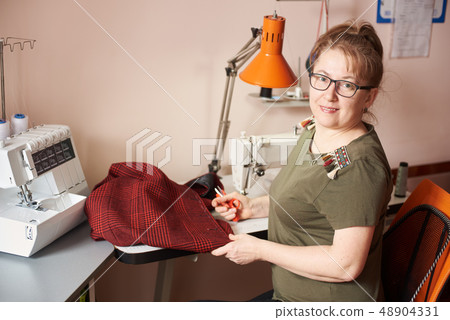 Smiling female tailor on sewing process of skirt, holding scissors. Overlock and sewing-machine on Smiling female tailor on sewing process of skirt, holding scissors. Overlock and sewing-machine on 48904331