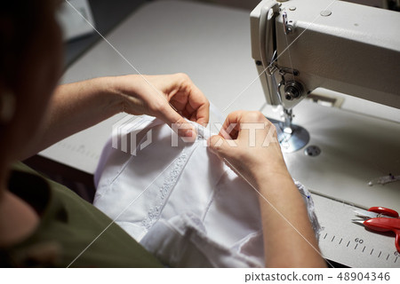 Needlewoman draping white garment with needle and her hands. Sewing machine, scissors on background 48904346