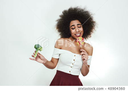 I love sweets so much! Cute afro american woman is facing a choice of healthy broccoli or lollipop 48904360