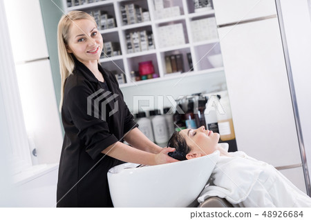 Smiling female blonde hairdresser looking at camera while working with client Smiling female blonde hairdresser looking at camera while working with client 48926684