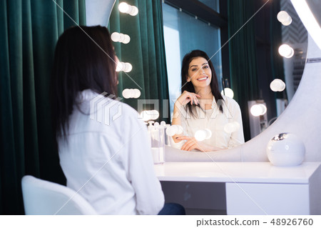 Smiling brunette woman looking at mirror while sitting at table indoors Smiling brunette woman looking at mirror while sitting at table indoors 48926760