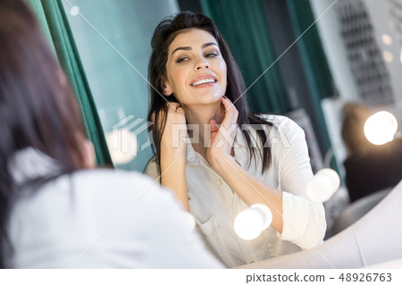 Beautiful woman in white bathrobe looking at mirror indoors Beautiful woman in white bathrobe looking at mirror indoors 48926763