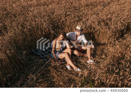 A young couple a man and a woman are sitting on a plaid, in summer in a wheat field. Relaxing on the A young couple a man and a woman are sitting on a plaid, in summer in a wheat field. Relaxing on the 48930800
