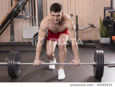 Young man flexing muscles with barbell in gym. Young man flexing muscles with barbell in gym. 48948424