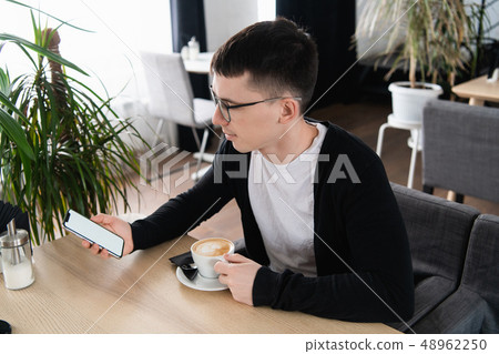 A photo of a handsome man wearing glasses looking at the screen of a mobile phone. Guy is sitting at 48962250