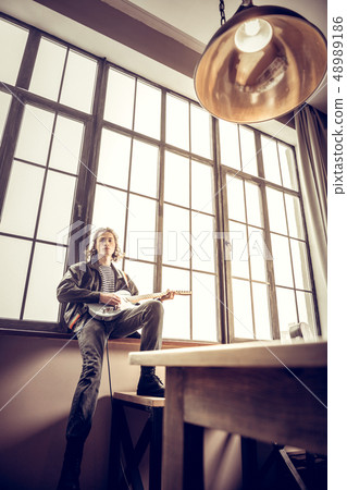 Slim rock musician wearing jeans sitting near window with guitar 48989186