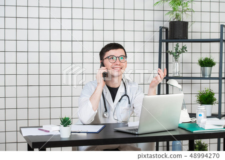 Male doctor using telephone while working on computer at table in clinic 48994270