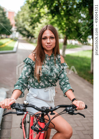 Girl in the summer in city, sitting on a bicycle, against the backdrop of trees and greenery. In a Girl in the summer in city, sitting on a bicycle, against the backdrop of trees and greenery. In a 49004331