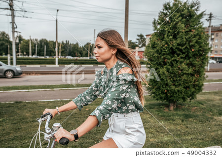 Girl in the summer in city, riding a bike, against the backdrop of trees and green cars. In a green Girl in the summer in city, riding a bike, against the backdrop of trees and green cars. In a green 49004332