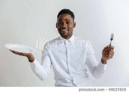 Young smiling attractive african american guy holding empty dish and fork isolated on grey 49025929