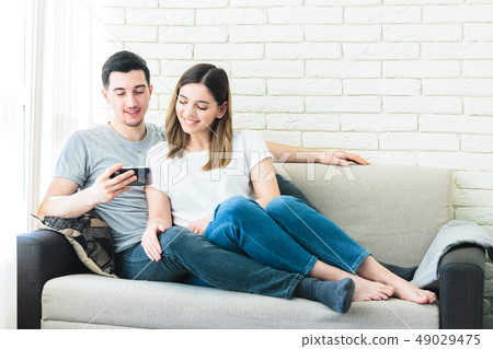 Relaxed couple using a generic mobile phone together sitting on a sofa in the living room at home 49029475