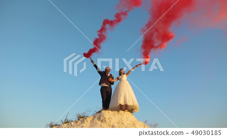 Happy bride and groom waving colored pink smoke against blue sky and laughing. Honeymoon. Romance 49030185