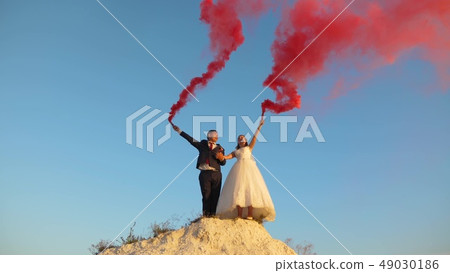 Happy bride and groom waving colored pink smoke against blue sky and laughing. Honeymoon. Romance Happy bride and groom waving colored pink smoke against blue sky and laughing. Honeymoon. Romance 49030186