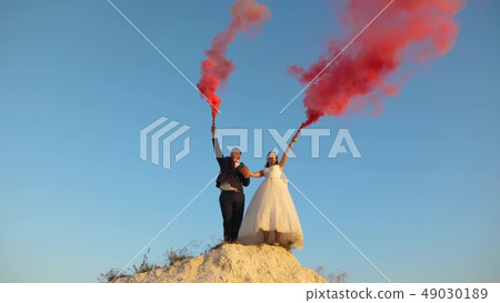 Happy bride and groom waving colored pink smoke against blue sky and laughing. Honeymoon. Romance 49030189