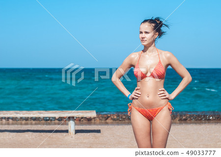 Beautiful and young girl in a striped orange swimsuite stands against the backdrop of the blue sea Beautiful and young girl in a striped orange swimsuite stands against the backdrop of the blue sea 49033777