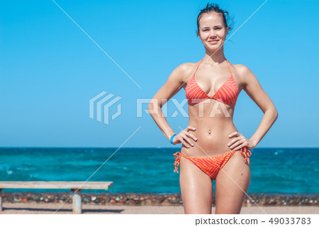 Beautiful and smiling young girl in a striped orange swimsuite stands against the backdrop of the Beautiful and smiling young girl in a striped orange swimsuite stands against the backdrop of the 49033783