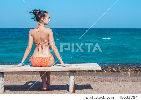 beautiful young girl in a striped orange swimsuite sits with her back to us on the bench over 49033784