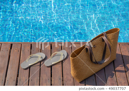 Flip flops and beach bag on wooden deck next to swimming pool. 49037707
