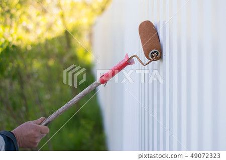 male hand paints a white metal fence with a roller 49072323