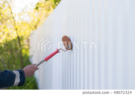 male hand paints a white metal fence with a roller 49072324
