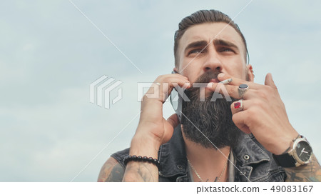 Brutal bearded man talking on the phone and smokes a cigarette. Brutal bearded man talking on the phone and smokes a cigarette. 49083167
