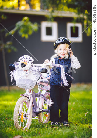 Funny child Caucasian girl blonde in a bicycle helmet near a purple bike with a basket in outside 49085886