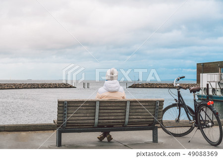 A young Caucasian woman sits with her back on a wooden bench overlooking the Baltic Sea on the A young Caucasian woman sits with her back on a wooden bench overlooking the Baltic Sea on the 49088369