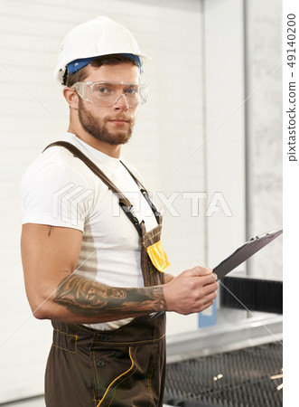 Brutal engineer in uniform holding folder, posing on factory Brutal engineer in uniform holding folder, posing on factory 49140200