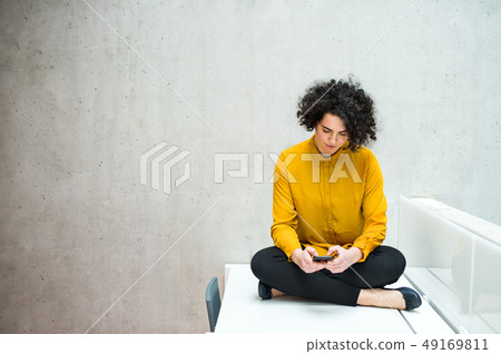 Young student or businesswoman with smartphone sitting on desk in room in a library or office. Young student or businesswoman with smartphone sitting on desk in room in a library or office. 49169811