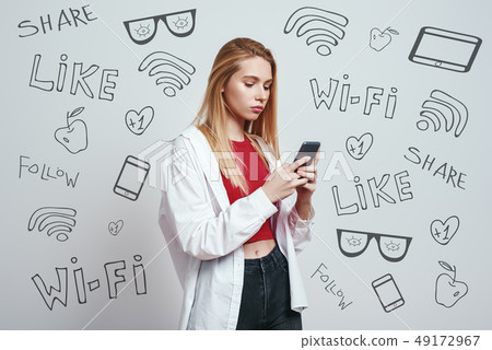 Surfing the net. Young blonde woman in a casual wear using her smart phone while standing against Surfing the net. Young blonde woman in a casual wear using her smart phone while standing against 49172967