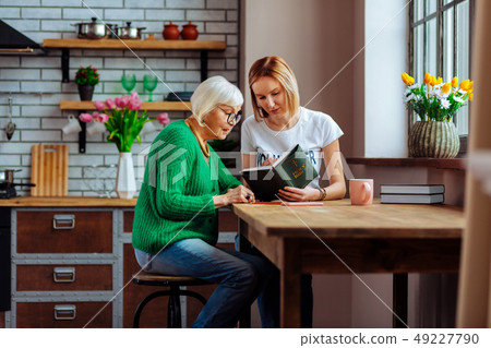 Young church volunteer reading Bible with pensioner at home kitchen 49227790