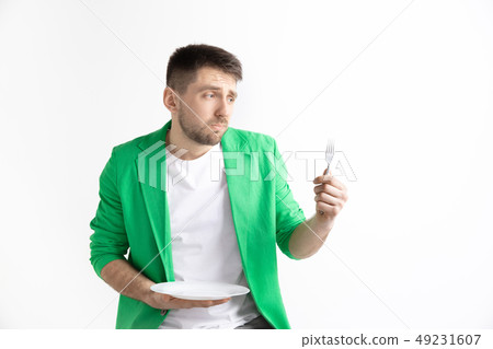 Young sad attractive guy holding empty dish and fork isolated on grey background. Young sad attractive guy holding empty dish and fork isolated on grey background. 49231607