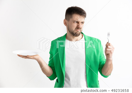 Young sad attractive guy holding empty dish and fork isolated on grey background. Young sad attractive guy holding empty dish and fork isolated on grey background. 49231658