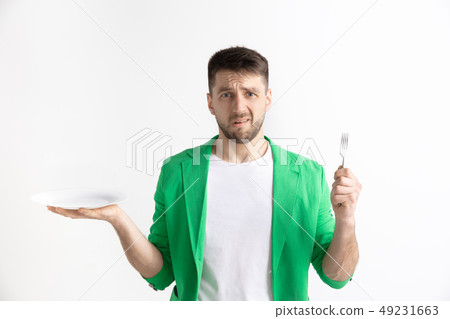 Young sad attractive guy holding empty dish and fork isolated on grey background. Young sad attractive guy holding empty dish and fork isolated on grey background. 49231663