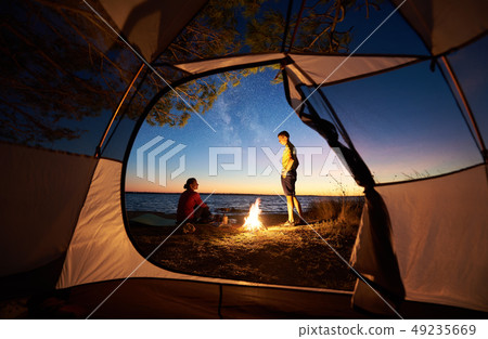 Young couple man and woman having rest at tourist tent and burning campfire on sea shore near forest Young couple man and woman having rest at tourist tent and burning campfire on sea shore near forest 49235669
