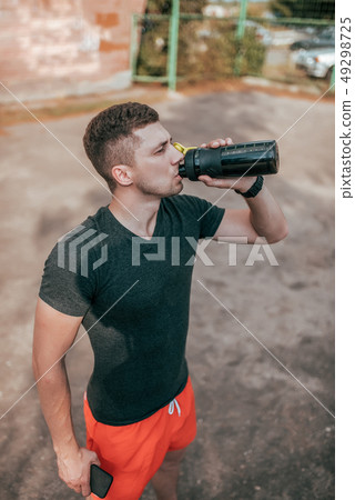 A male athlete, in the summer in the city on sports field, drinks clean water from a bottle shaker 49298725